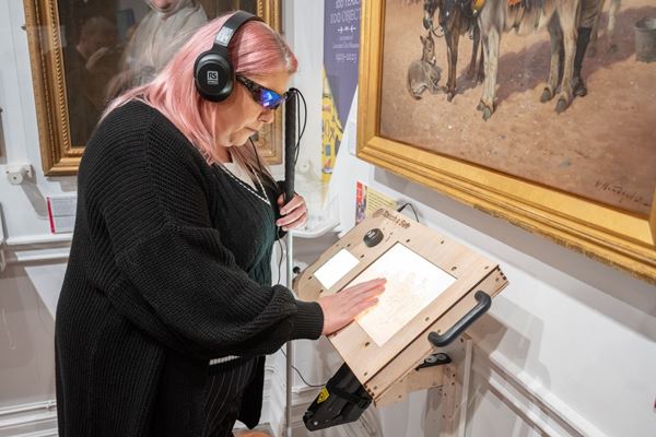 Photo of a woman wearing headphones and exploring the Touch & See lithophane recreation of the painting 'Donkey Boy', which depicts a group of donkeys on Morecambe beach with the young man looking after them..